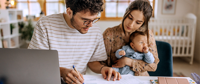 Father and mother holding their newborn baby reviewing documents in front of their laptop.