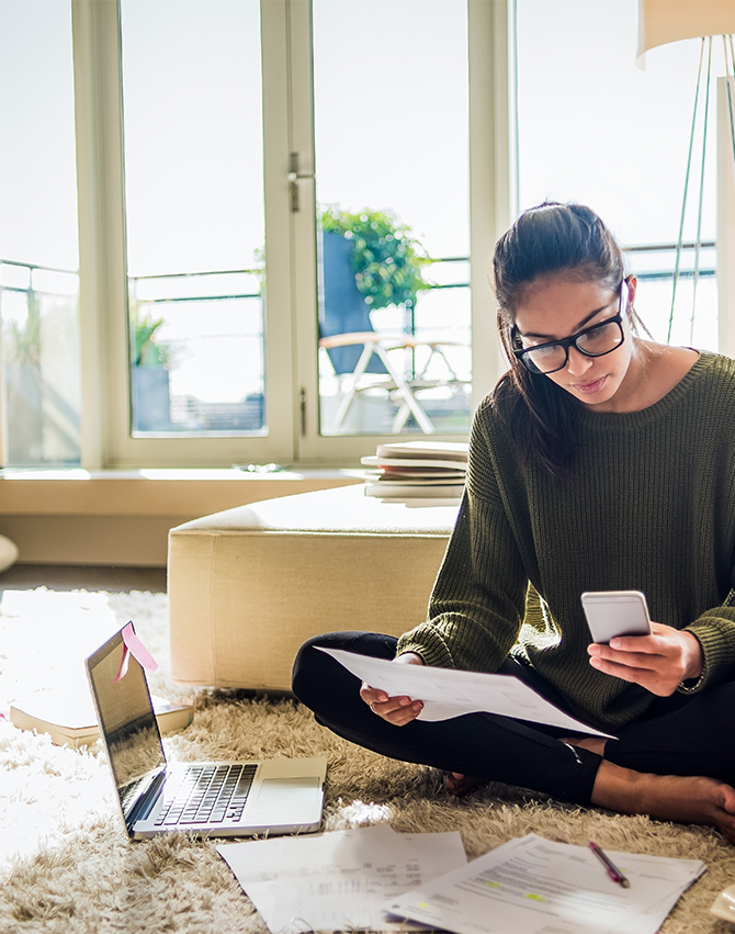 Woman sitting on the floor of a living room reviewing financial documents next to an open laptop.